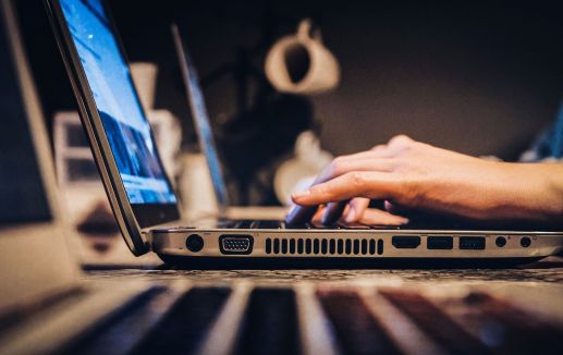 Eine Person tippt auf einer Computertastatur. Der Computer steht auf einem Tisch. Im Vordergrund und Hintergrund sind weitere Laptops verschwommen zu erkennen.