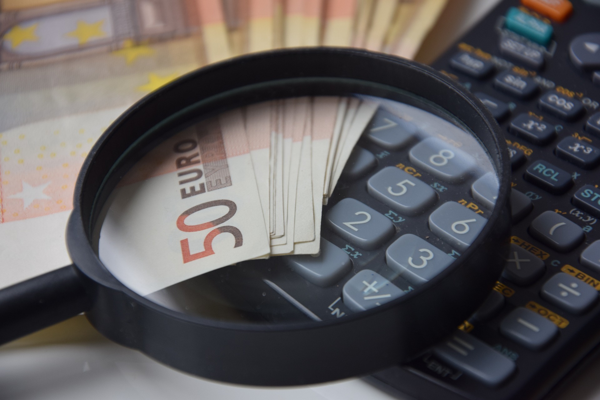 Eine Lupe vergrößert mehrere 50-Euro-Scheine, die auf einem Taschenrechner liegen. Im Hintergrund sind weitere Euro-Scheine sichtbar, was einen Finanz- oder Rechnungszusammenhang andeutet.