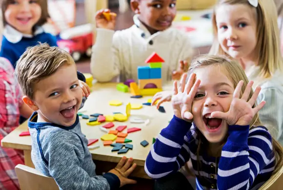 Eine Gruppe Kindergartenkinder sitze an einem Tisch in einem Klassenzimmer. Sie spielen  mit bunten Holzblöcken. Im Vordergrund sitzt ein Mädchen und macht eine Grimasse.
