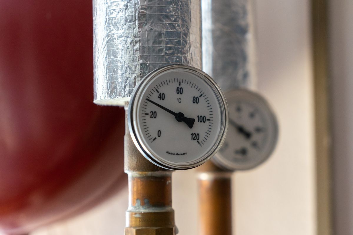 Ein Temperaturmessgerät zeigt etwa 60 Grad Celsius an, montiert auf einem isolierten Rohr. Im Hintergrund ist ein weiteres Messgerät leicht unscharf zu erkennen.