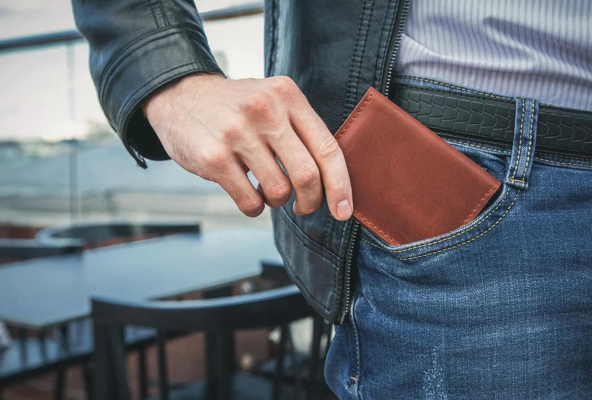 Eine Hand steckt ein braunes Portemonnaie in die Hosentasche einer blauen Jeans. Im Hintergrund sind unscharfe Tische und Stühle auf einer Terrasse erkennbar.