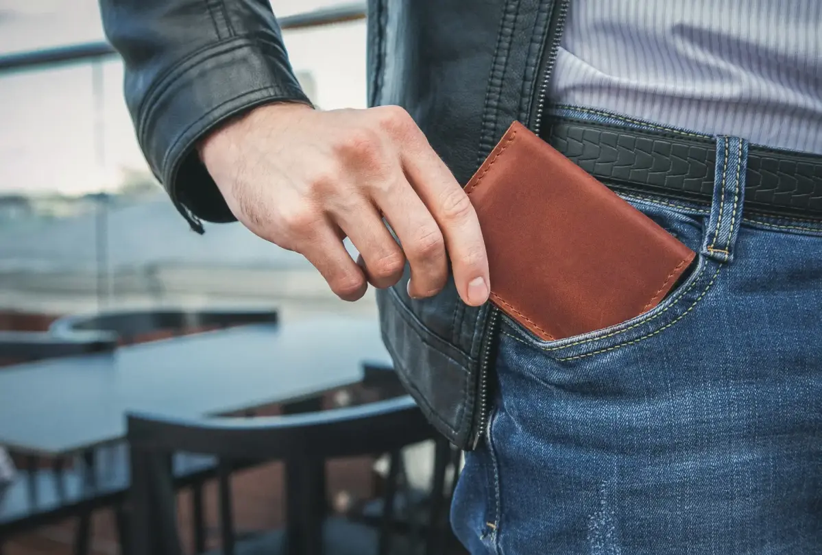 Eine Hand steckt ein braunes Portemonnaie in die Hosentasche einer blauen Jeans. Im Hintergrund sind unscharfe Tische und Stühle auf einer Terrasse erkennbar.