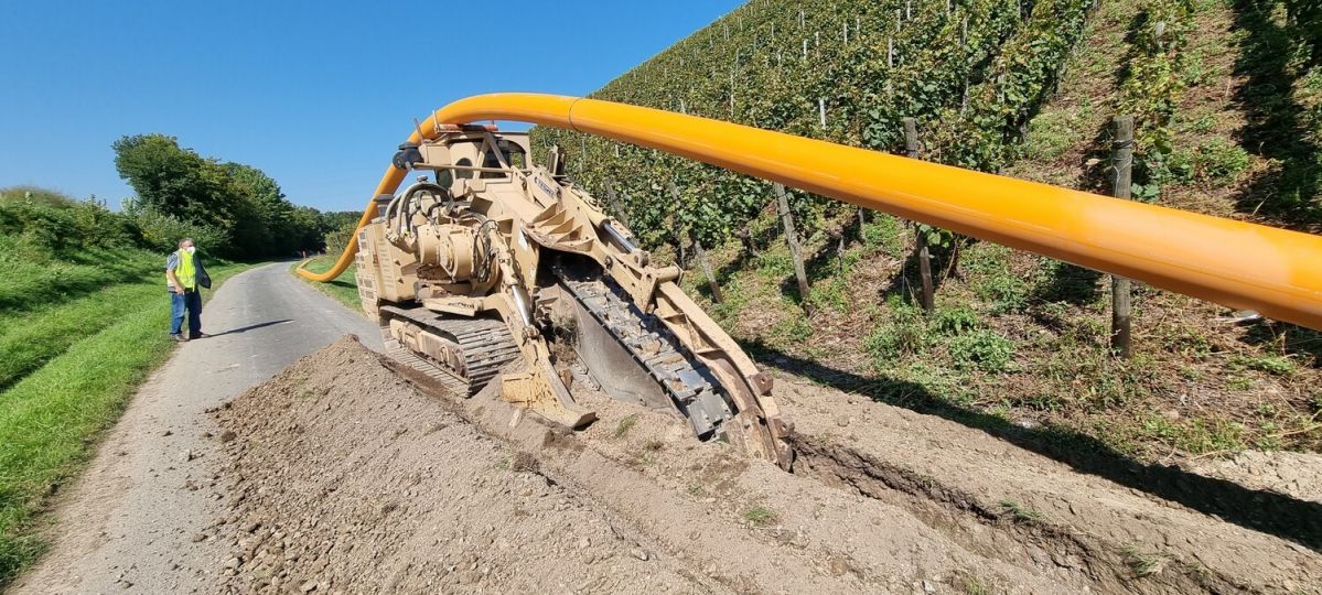 Eine große, gelbe Baumaschine gräbt einen Graben entlang einer Straße. Ein Arbeiter steht daneben. Im Hintergrund sind Weinreben auf einem Hügel zu sehen unter einem klaren, blauen Himmel.