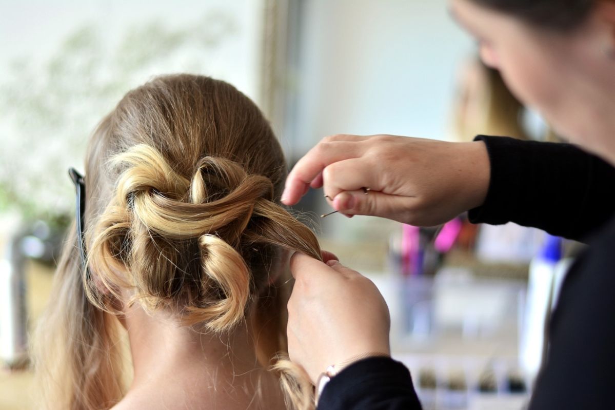 Für Schönheit und Wohlbefinden Eine Frisur wird von einer Person gestaltet, die mit den Händen sorgfältig Locken in einem eleganten Knoten formt. Im Hintergrund sind verschwommen Friseurwerkzeuge zu sehen.
