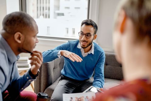 Drei professionell gekleidete Männer sitzen auf einem grauen Sofa und diskutieren miteinander. 