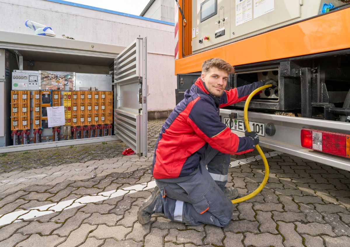 Ein Mann kniet auf einem gepflasterten Boden, verbindet ein gelbes Kabel mit einem orangefarbenen Generatorfahrzeug. Im Hintergrund steht ein offenes Schaltkastenmodell mit mehreren Anschlüssen.
