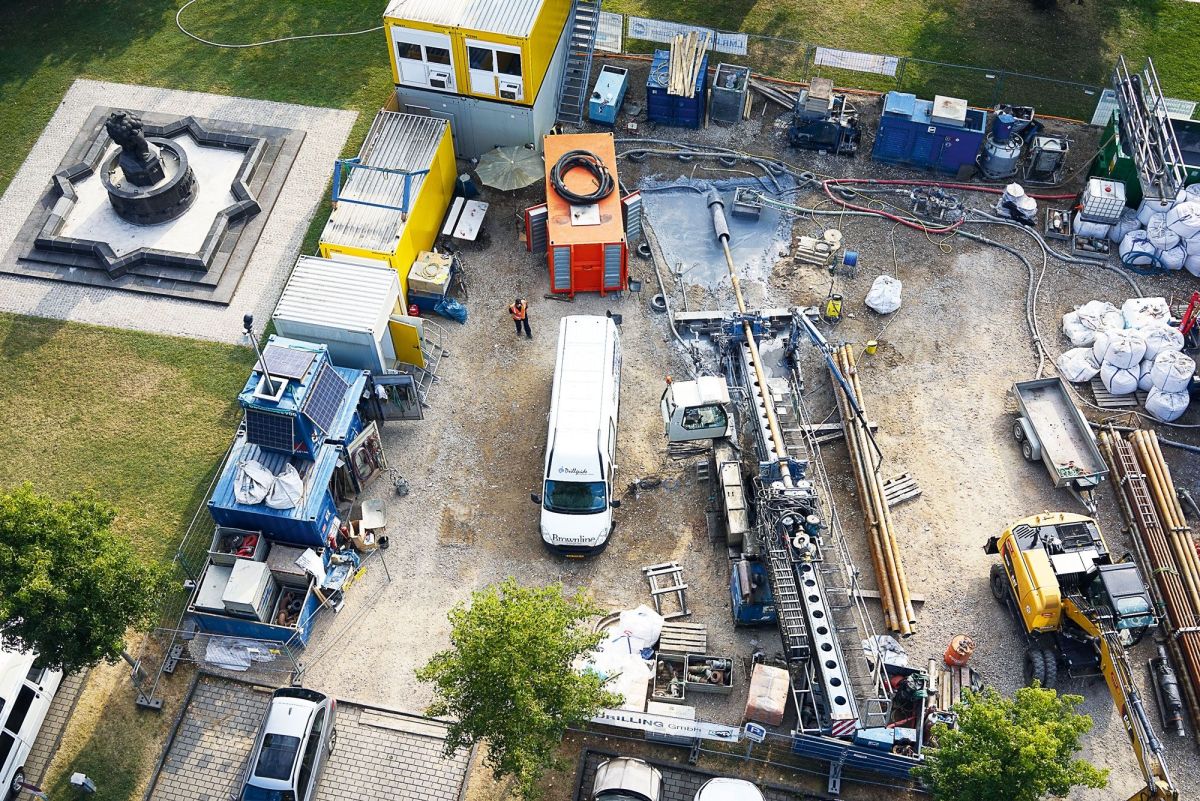 Aufnahme von oben auf eine Baustelle. Auf der Baustelle befinden sich Container, ein Bagger, ein Transporter, Rohre und eine Person in Schutzkleidung.