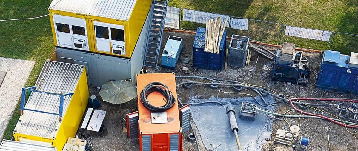 Aufnahme von oben auf eine Baustelle. Auf der Baustelle befinden sich Container, ein Bagger, ein Transporter, Rohre und eine Person in Schutzkleidung.