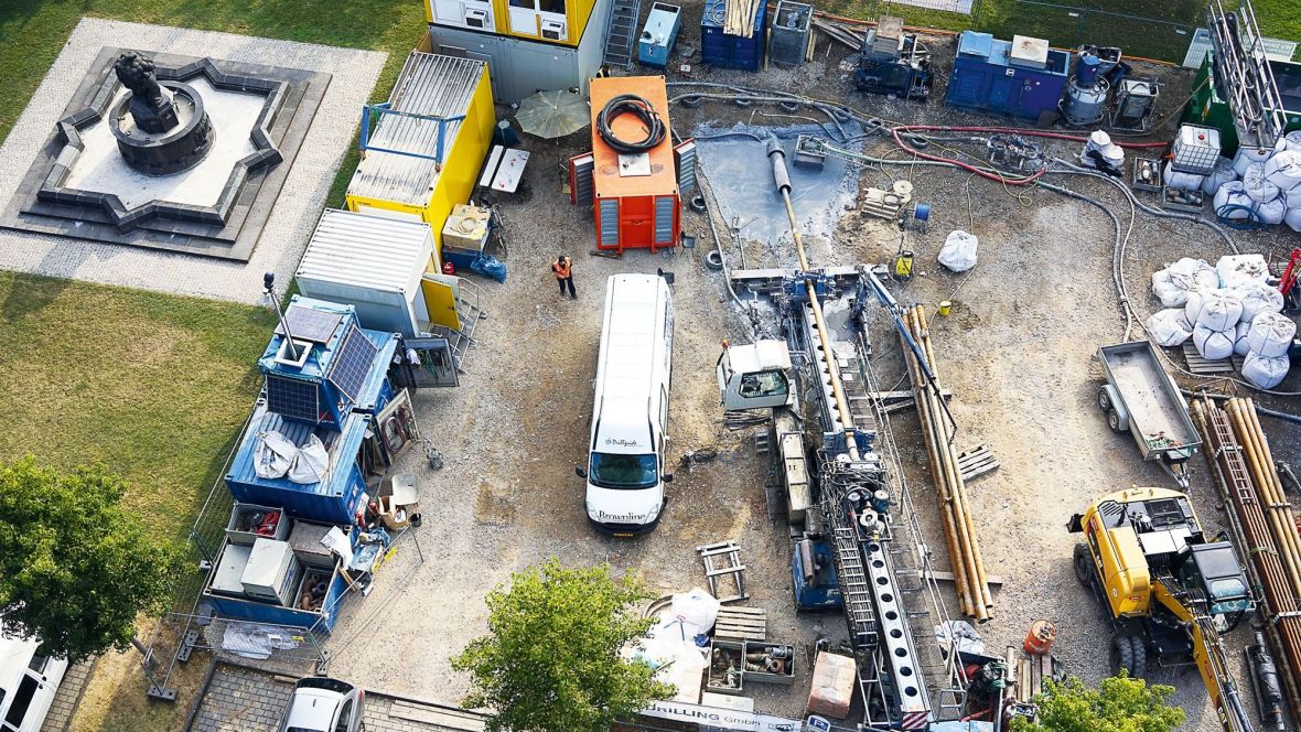 Aufnahme von oben auf eine Baustelle. Auf der Baustelle befinden sich Container, ein Bagger, ein Transporter, Rohre und eine Person in Schutzkleidung.
