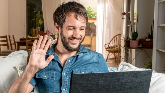 Ein Mann sitzt auf einem Sofa, lächelt und winkt einem Laptop zu. Im Hintergrund befindet sich ein gemütlicher, heller Raum mit Pflanzen und Holzmöbeln.