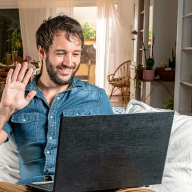 Ein Mann sitzt auf einem Sofa, lächelt und winkt einem Laptop zu. Im Hintergrund befindet sich ein gemütlicher, heller Raum mit Pflanzen und Holzmöbeln.