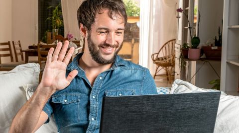 Ein Mann sitzt auf einem Sofa, lächelt und winkt einem Laptop zu. Im Hintergrund befindet sich ein gemütlicher, heller Raum mit Pflanzen und Holzmöbeln.