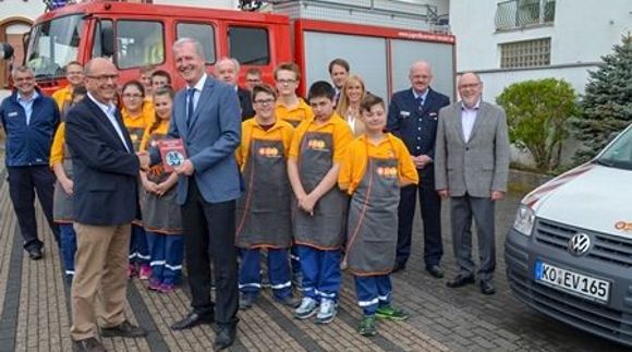 Eine Gruppe an Kindern in evm Schürzen und Feuerwehr Uniform steht vor einem Feuerwehrauto. Neben ihnen stehen mehrere professionell gekleidete Erwachsene Personen.