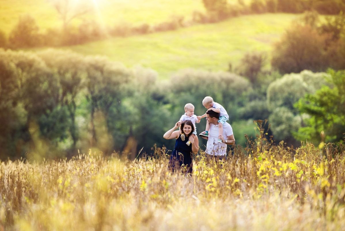 Eine Familie spaziert durch eine blühende Wiese, während ein Kind auf den Schultern des Vaters sitzt. Im Hintergrund sind Bäume und ein Hügel in Sonnenlicht zu sehen.