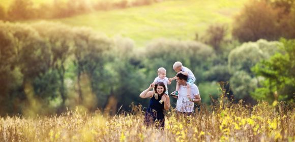 Eine Familie spaziert durch eine blühende Wiese, während ein Kind auf den Schultern des Vaters sitzt. Im Hintergrund sind Bäume und ein Hügel in Sonnenlicht zu sehen.