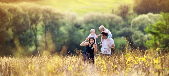 Eine Familie spaziert durch eine blühende Wiese, während ein Kind auf den Schultern des Vaters sitzt. Im Hintergrund sind Bäume und ein Hügel in Sonnenlicht zu sehen.
