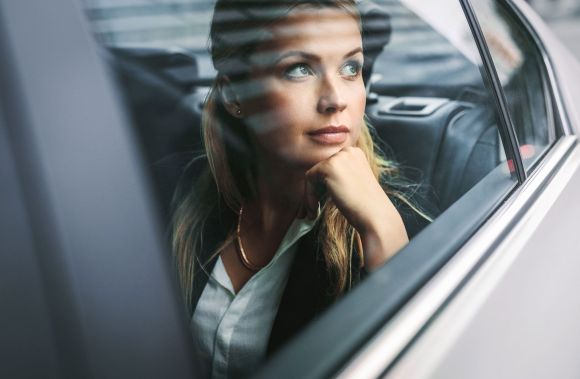 Eine Frau sitzt nachdenklich im Auto, den Kopf auf ihre Hand gestützt, und blickt aus dem Fenster. Draußen sind schemenhafte Strukturen eines urbanen Umfelds erkennbar.