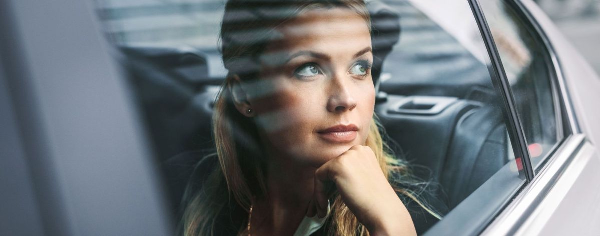 Eine Frau sitzt nachdenklich im Auto, den Kopf auf ihre Hand gestützt, und blickt aus dem Fenster. Draußen sind schemenhafte Strukturen eines urbanen Umfelds erkennbar.