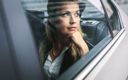 Eine Frau sitzt nachdenklich im Auto, den Kopf auf ihre Hand gestützt, und blickt aus dem Fenster. Draußen sind schemenhafte Strukturen eines urbanen Umfelds erkennbar.