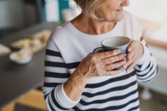 Eine ältere Frau hält eine graue Tasse in beiden Händen, lächelt leicht und trägt einen gestreiften Pullover. Im Hintergrund ist ein unscharfer Tisch zu sehen.