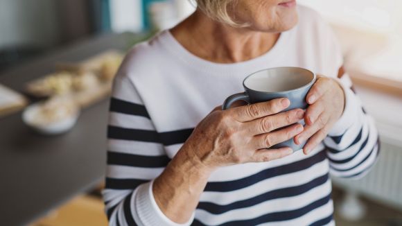 Eine ältere Frau hält eine graue Tasse in beiden Händen, lächelt leicht und trägt einen gestreiften Pullover. Im Hintergrund ist ein unscharfer Tisch zu sehen.