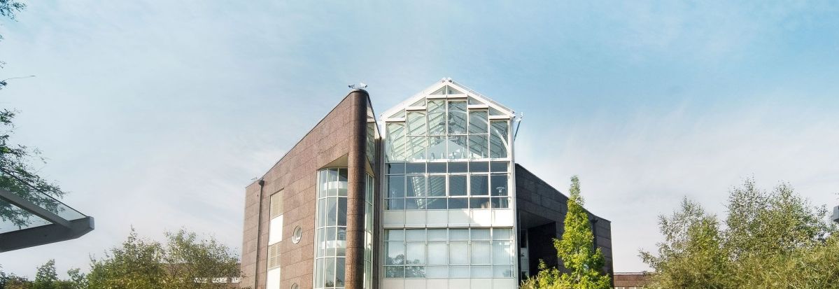 Ein modernes Gebäude mit einem großen Glasdach steht im Zentrum. Ein betonierter Weg führt dorthin, umgeben von grüner Vegetation unter einem blauen Himmel.