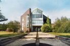 Ein modernes Gebäude mit einem großen Glasdach steht im Zentrum. Ein betonierter Weg führt dorthin, umgeben von grüner Vegetation unter einem blauen Himmel.