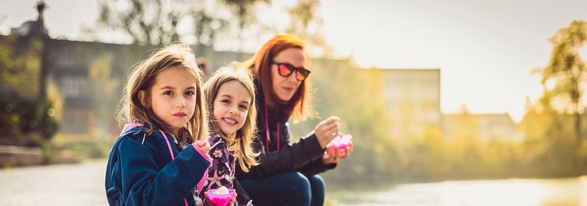 Eine Frau sitzt mit 2 Mädchen am Ufer eines Flusses. Sie haben Eisbecher in der Hand und lächeln freundlich.
