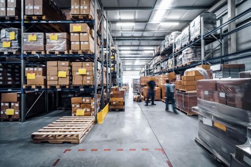 Eine große Lagerhalle mit Paketen. Zwei Personen sind verschwommen bei der Arbeit zu sehen.