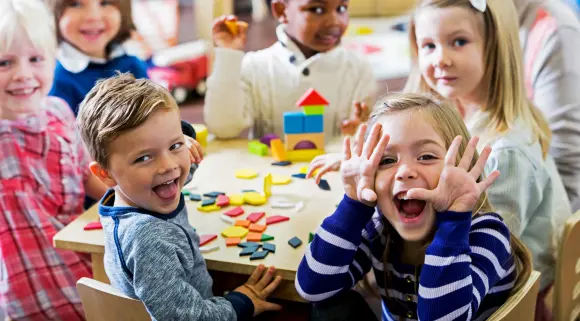 Eine Gruppe Kindergartenkinder sitze an einem Tisch in einem Klassenzimmer. Sie spielen  mit bunten Holzblöcken. Im Vordergrund sitzt ein Mädchen und macht eine Grimasse.