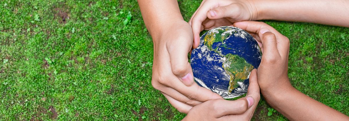 Vier Hände sind um eine kleine Weltkugel umschlungen. Im Hintergrund ist ein Rasen zu erkennen.
