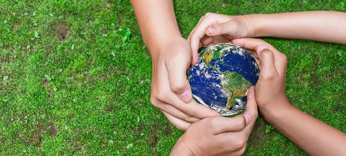 Vier Hände sind um eine kleine Weltkugel umschlungen. Im Hintergrund ist ein Rasen zu erkennen.
