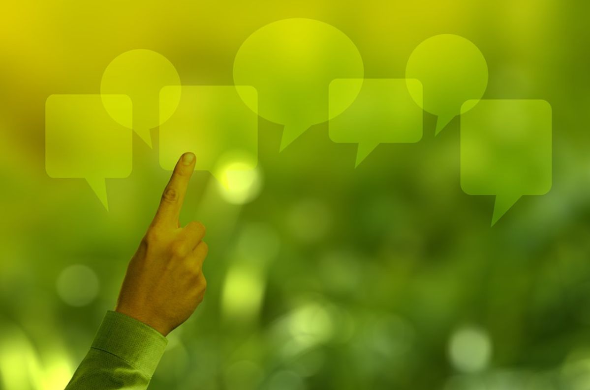 Eine Hand zeigt auf eine Reihe grüner Sprechblasen, die im Vordergrund schweben. Im Hintergrund ist eine verschwommene, natürliche Grünlandschaft zu sehen.