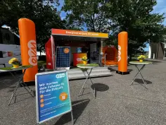 Das orangene Promomobil der evm steht auf einem Platz. Drei Stehtische stehen davor und eine blaue Werbetafel. Neben dem Mobil stehen 2 orangene evm Pfeiler.