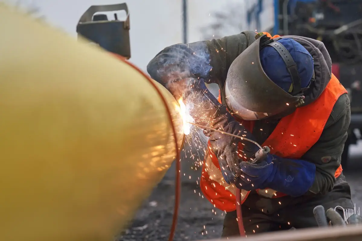 Eine Person mit Schutzkleidung schweißt etwas an einem großen gelben Rohr.