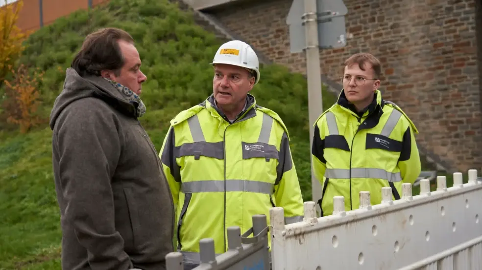 Zwei Männer in gelben Schutzjacken und weißen Schutzhelmen stehen an einer Absperrung und reden mit einem normal gekleideten Mann.