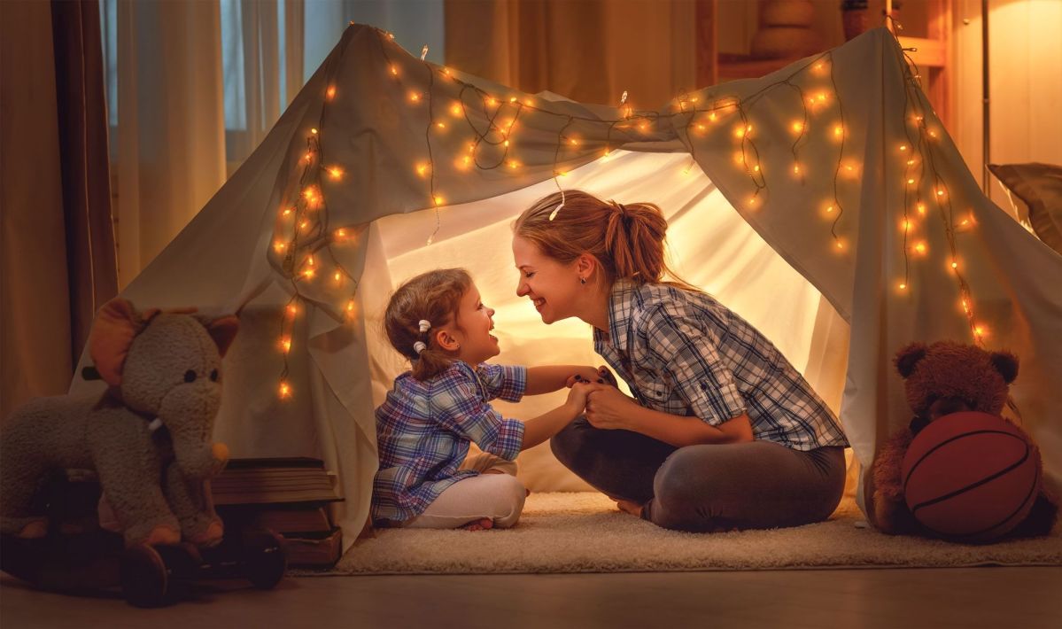 Eine Mutter und ihr Kind sitzen lächelnd in einem mit Lichterketten geschmückten, selbstgebauten Zelthaus im Wohnzimmer, umgeben von Kuscheltieren und einem Basketball.