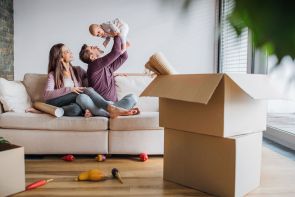 Ein Mann hält lachend ein Baby in die Luft. Eine Frau sitzt daneben auf dem Sofa. Im Vordergrund steht ein geöffneter Umzugskarton mit Decken.