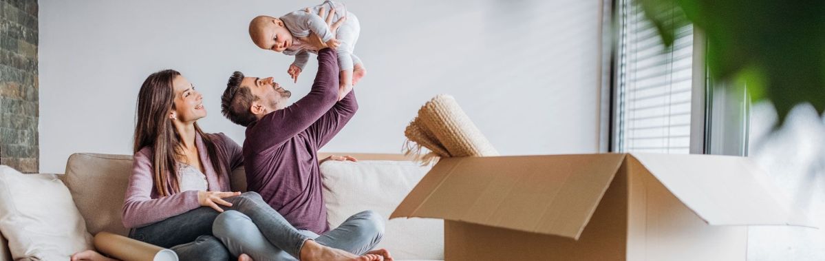 Ein Mann hält lachend ein Baby in die Luft. Eine Frau sitzt daneben auf dem Sofa. Im Vordergrund steht ein geöffneter Umzugskarton mit Decken.