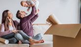 Ein Mann hält lachend ein Baby in die Luft. Eine Frau sitzt daneben auf dem Sofa. Im Vordergrund steht ein geöffneter Umzugskarton mit Decken.