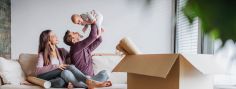 Ein Mann hält lachend ein Baby in die Luft. Eine Frau sitzt daneben auf dem Sofa. Im Vordergrund steht ein geöffneter Umzugskarton mit Decken.
