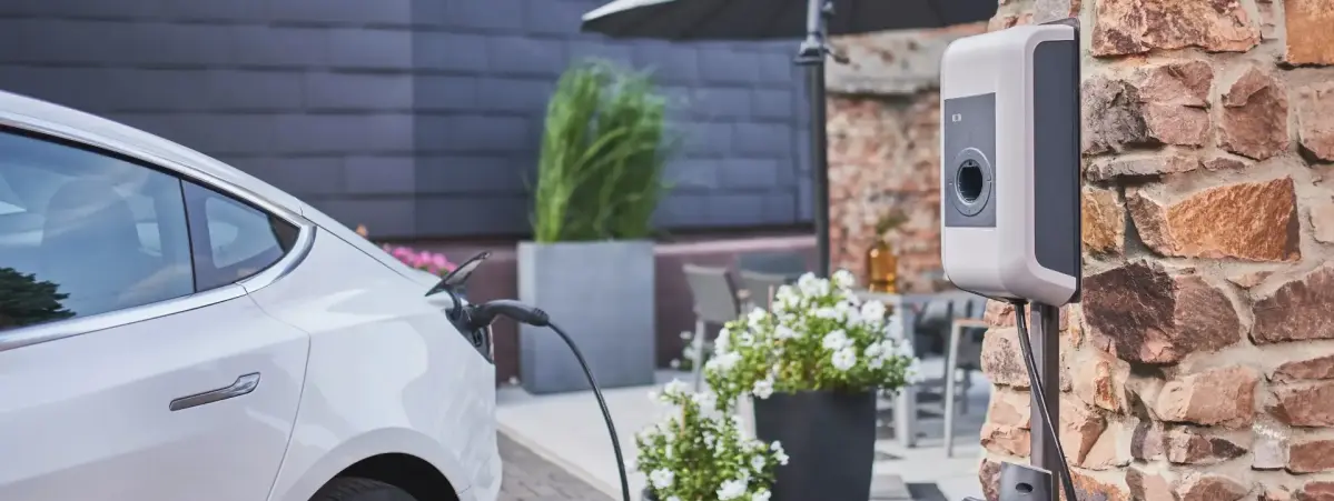 Ein E-Auto lädt an einer Wallbox die an einer Hauswand angebracht ist. Im Hintergrund ist ein Tisch und ein Sonnenschirm zu erkennen.