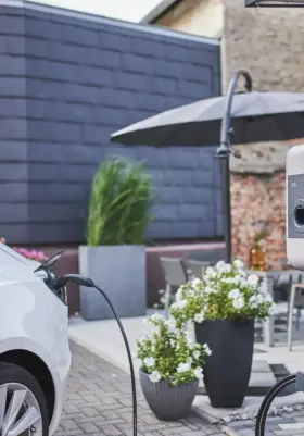 Ein E-Auto lädt an einer Wallbox die an einer Hauswand angebracht ist. Im Hintergrund ist ein Tisch und ein Sonnenschirm zu erkennen.