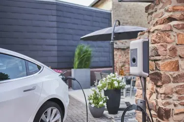 Ein E-Auto lädt an einer Wallbox die an einer Hauswand angebracht ist. Im Hintergrund ist ein Tisch und ein Sonnenschirm zu erkennen.