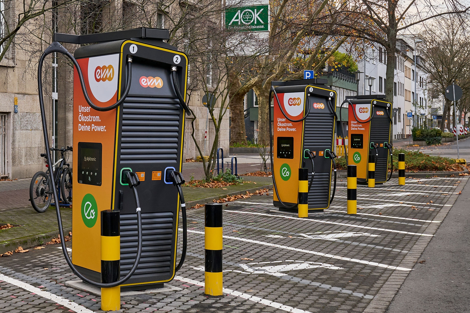 Drei Ladesäulen der evm in Orange und Schwarz auf leeren Parkplätzen mit E-Auto-Symbol