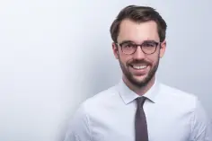 Ein professionell gekleideter Mann mit dunklen Haaren und einer Brille lächelt freundlich.
