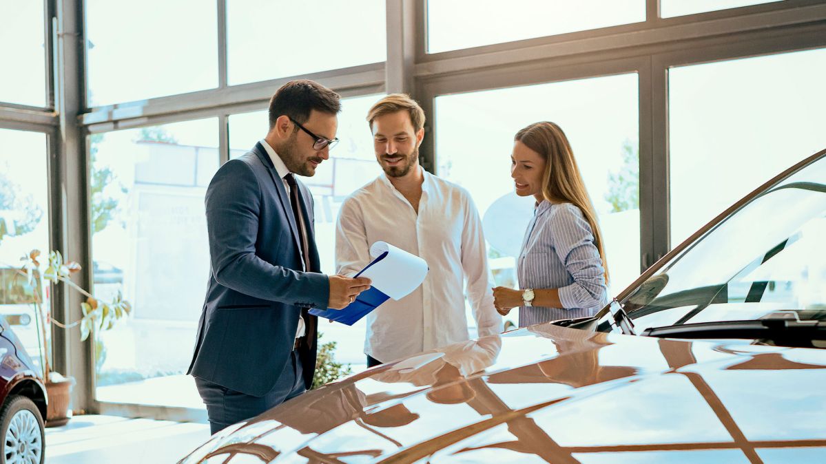 Ein professionell gekleideter Mann mit einem clipboard zeigt einem Paar etwas. Sie stehen vor einem Auto in einem Autohaus.
