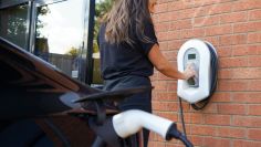 Eine Frau bedient eine Ladestation, die an einer roten Ziegelmauer befestigt ist. Ein Elektroauto ist im Vordergrund angeschlossen. Rechts befindet sich ein großes Fenster.