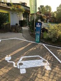 Ein E-Auto Icon ist weiß gesprüht auf einem Parkplatz. Vor dem Parkplatz steht eine blaue evm Ladesäule.