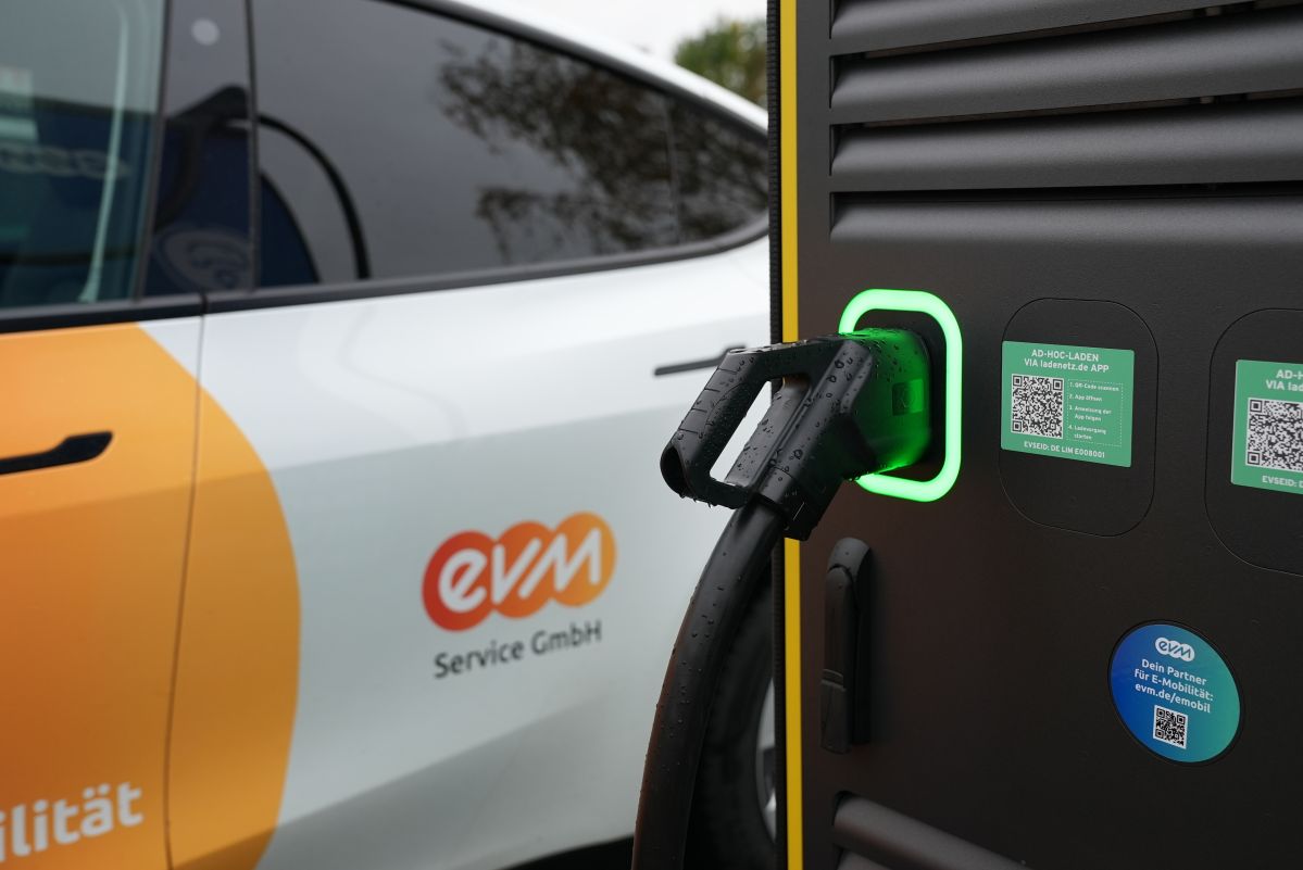 Ladeinfrastruktur Ein Elektroauto wird an eine Ladestation angeschlossen; der Ladenstecker steckt im Auto. Die Haltestation zeigt leuchtende Symbole und QR-Codes. Das Auto trägt das Logo „evm Service GmbH“.